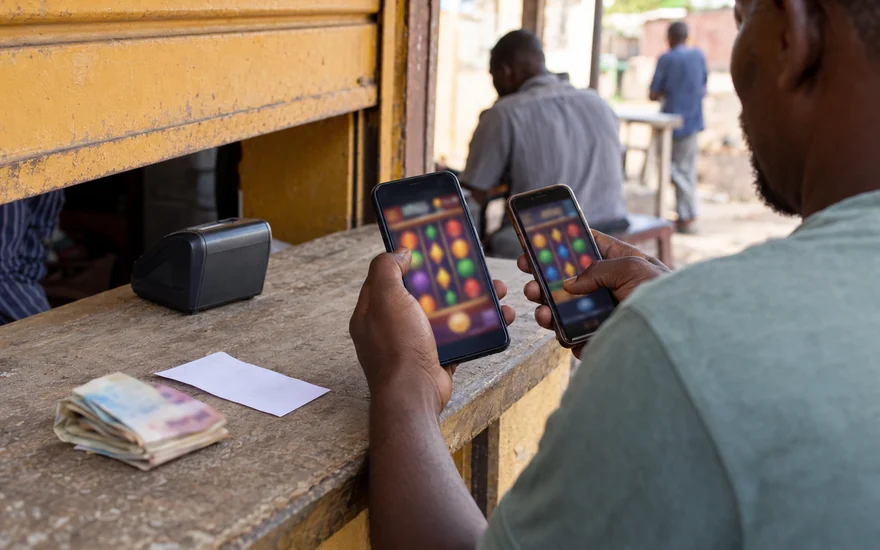 A 5 dollar deposit casino comparison illustrated by two blurred casino phones, anonymous folded cash and a blank betting coupon at a Lusaka kiosk.