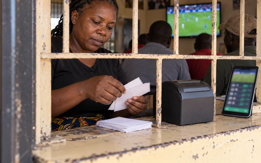 GSB ticket check illustrated by a betting-shop cashier sorting anonymous coupons beside a terminal and blurred betting phone.