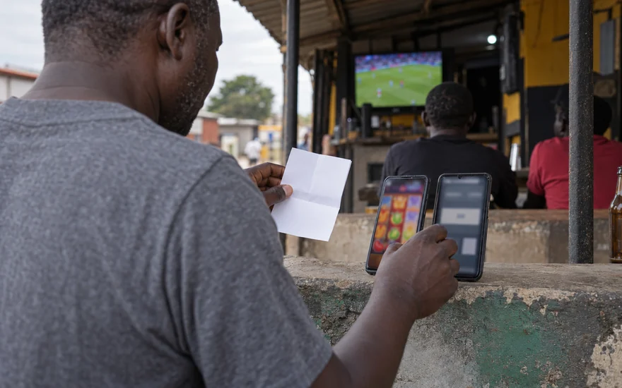 Crypto casino comparison for Zambia comparison scene with two blurred phones, a betting coupon and a distant football screen outside a local betting bar.